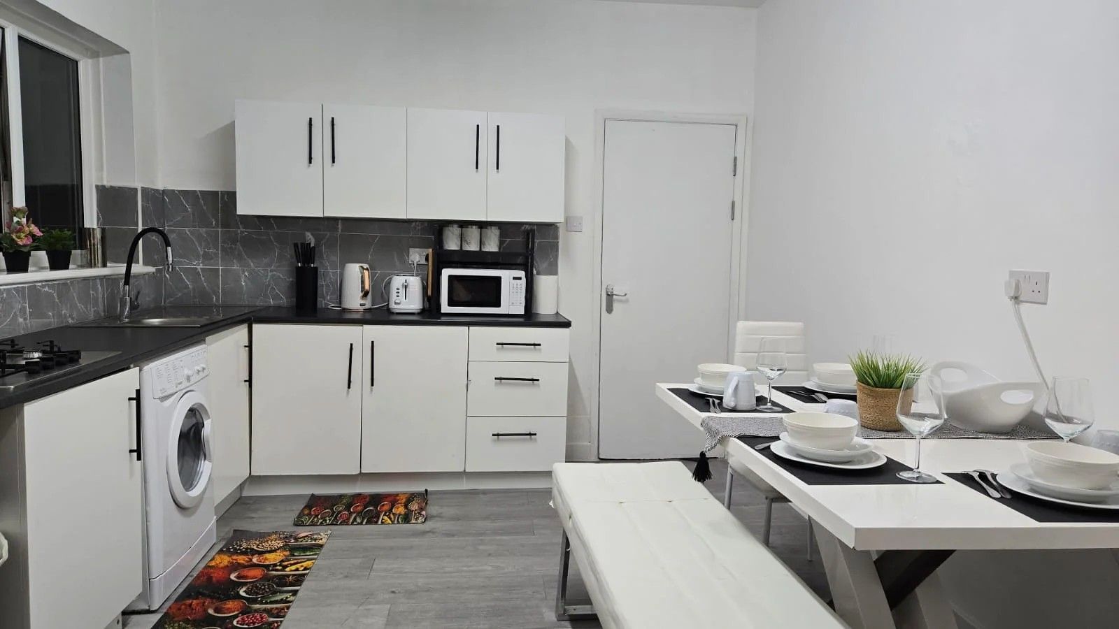A modern kitchen with white cabinets, a built-in washing machine, a microwave, and a dining area with a bench and table set for two.