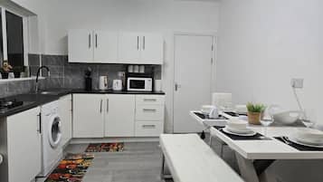 A modern kitchen with white cabinets, a built-in washing machine, a microwave, and a dining area with a bench and table set for two.