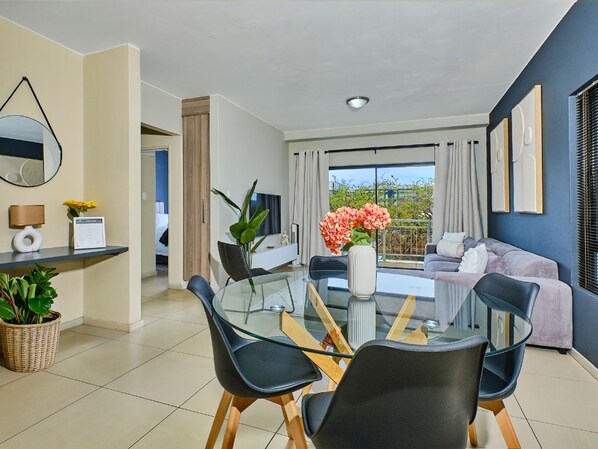 A modern living room with a glass dining table, black chairs, a sofa, and a large window with curtains. - Regentis Executive Stays at The Atrium Unit 164 (Johannesburg)