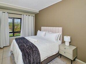A bedroom with a bed, a nightstand, a window with curtains, and a view outside. - Regentis Executive Stays at The Atrium Unit 164 (Johannesburg)