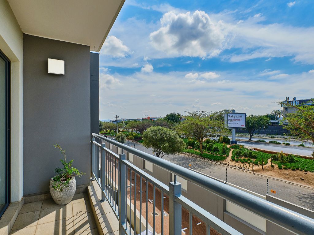 Een balkon met een bloempot, een metalen hekwerk en uitzicht op een weg met groen en een gebouw in de verte.