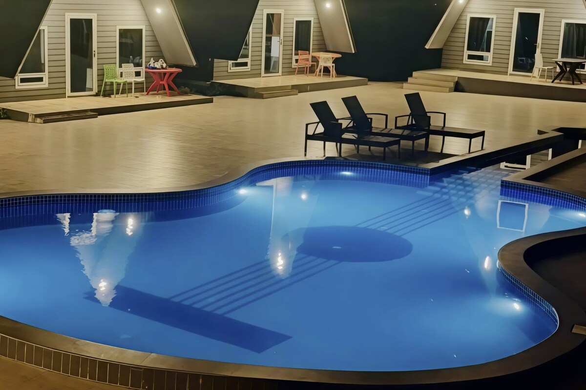 Una piscina con sillones de descanso cercanos y una mesa pequeña, rodeada por un edificio de techo triangular y varias ventanas.