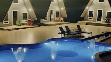 A swimming pool with adjacent lounge chairs and a small table, surrounded by a building with a triangular roof and multiple windows.