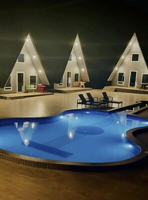 Una piscina con sillones de descanso cercanos y una mesa pequeña, rodeada por un edificio de techo triangular y varias ventanas.