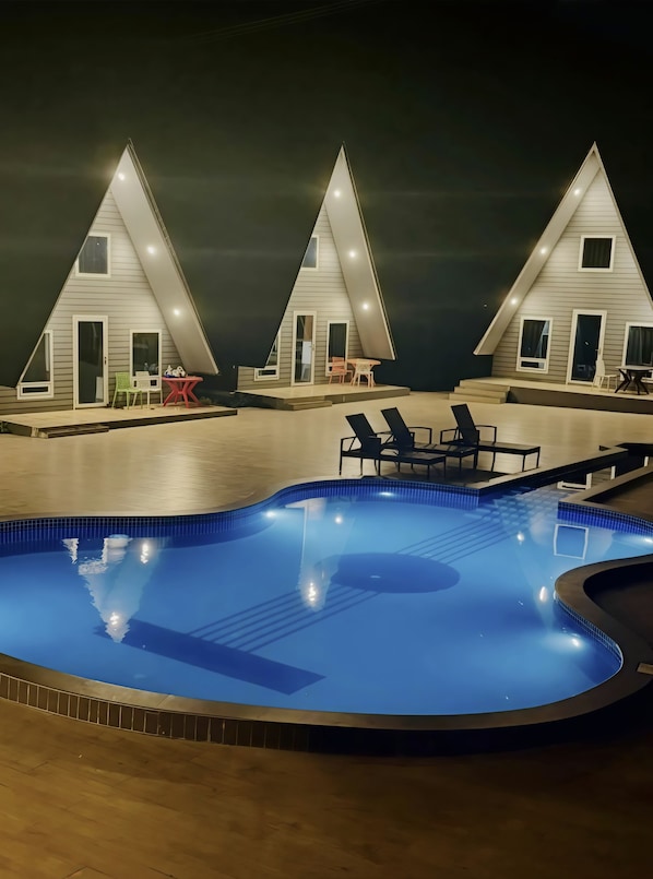 A swimming pool with adjacent lounge chairs and a small table, surrounded by a building with a triangular roof and multiple windows.