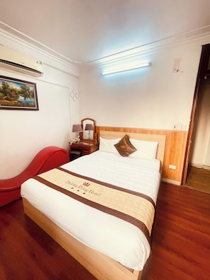Una habitación de hotel con una cama, una mesita de noche, una lámpara, una silla roja, un cuadro en la pared y un cabecero de madera.