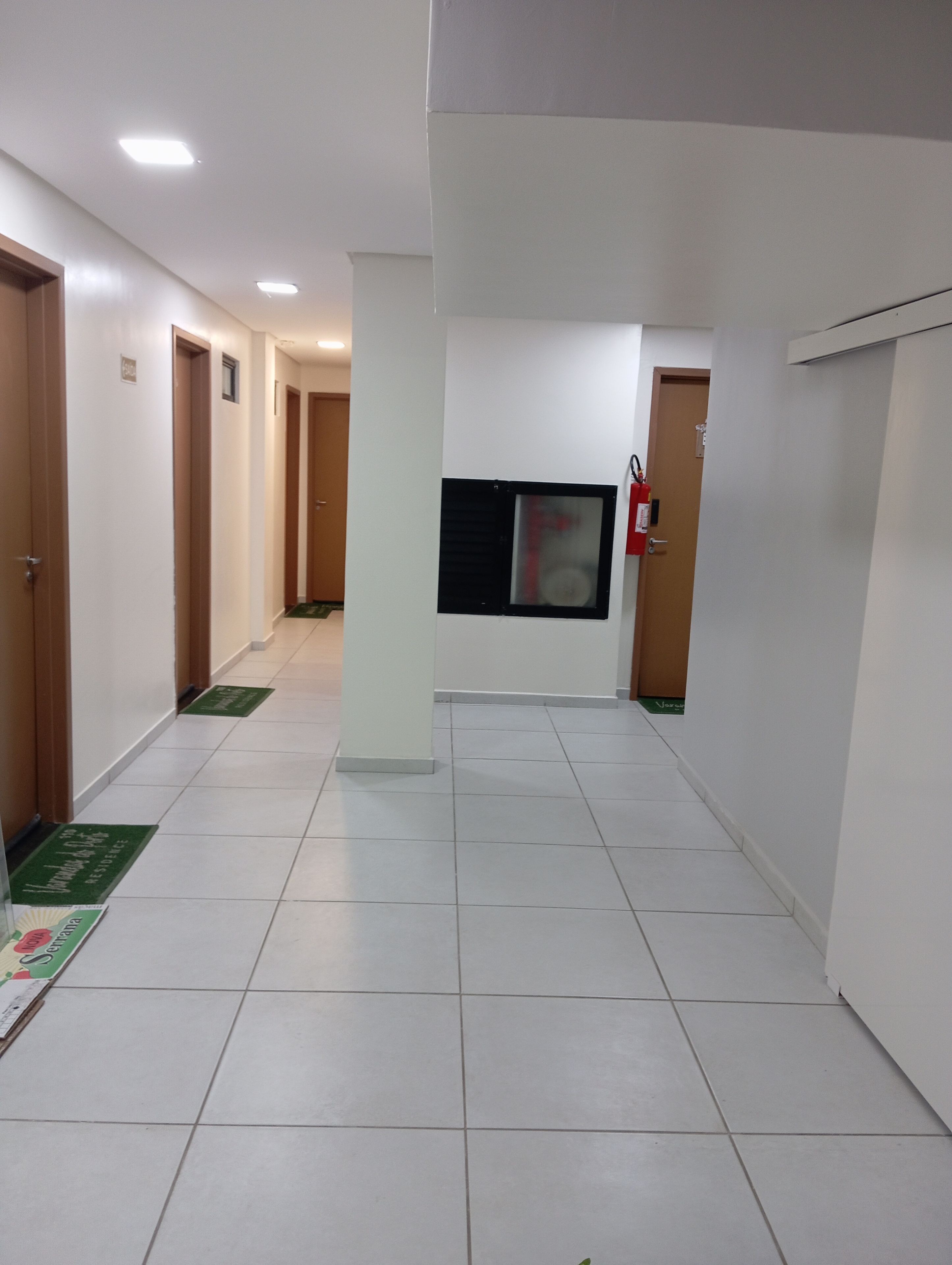 Un pasillo con pisos de baldosas blancas, puertas de madera y un extintor colgado en la pared.