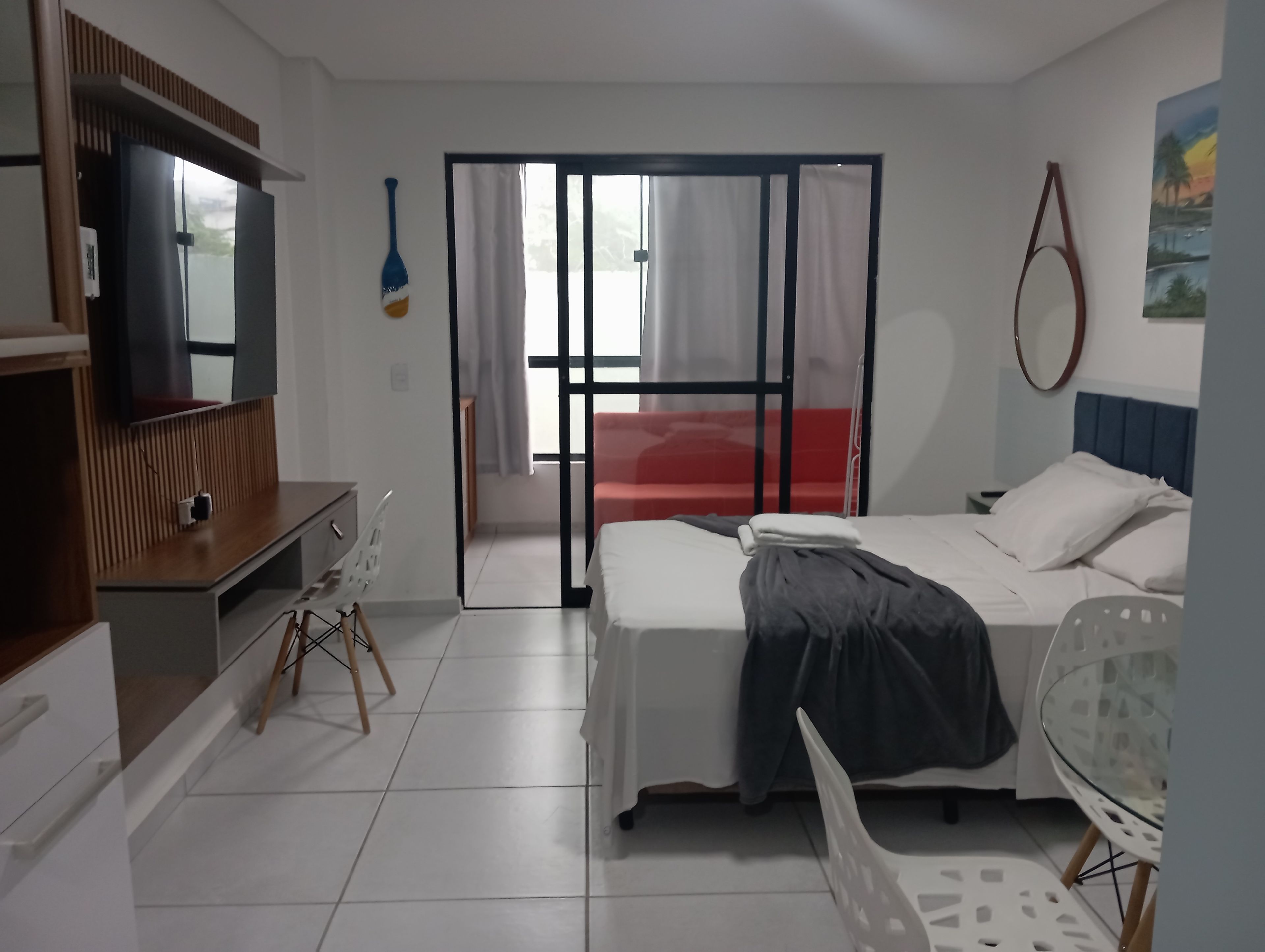 Habitación de hotel con cama, escritorio, silla, televisor y puerta corrediza a un balcón.