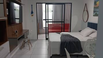 Habitación de hotel con cama, escritorio, silla, televisor y puerta corrediza a un balcón.