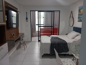 Habitación de hotel con cama, escritorio, silla, televisor y puerta corrediza a un balcón.