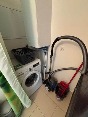Un espace dédié à la lessive, comprenant une machine à laver, un panier à linge, un aspirateur et une serpillière.