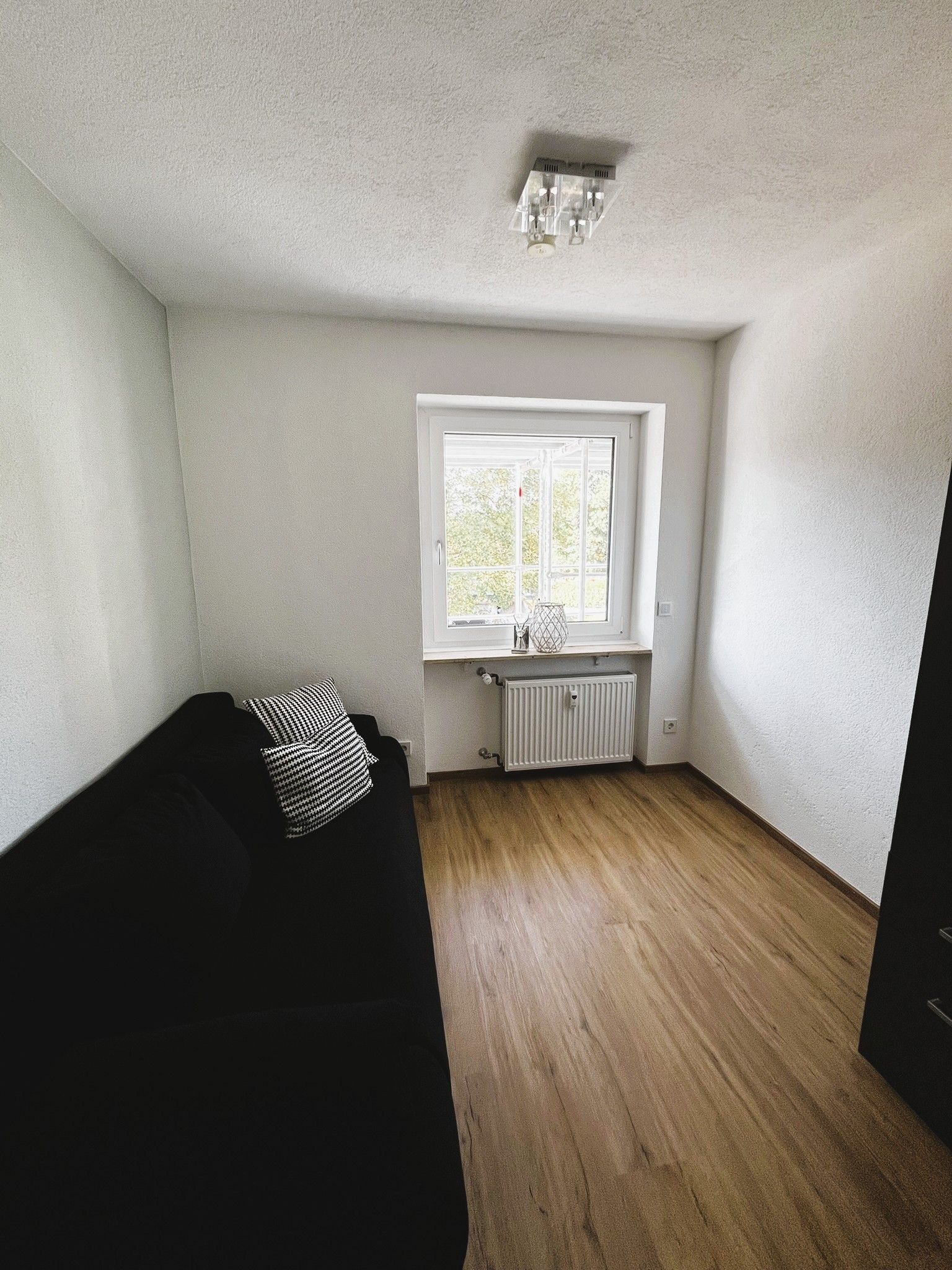 Ein Zimmer mit einer schwarzen Couch, einem Fenster mit Blick, einer Heizung und einer Deckenleuchte.