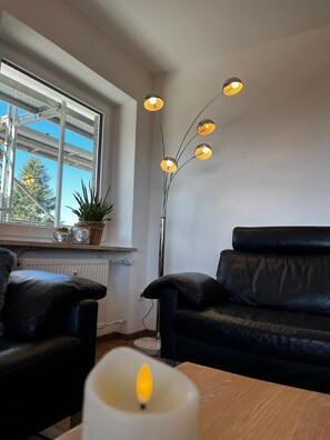 A room with a black leather sofa, a white table with a lit candle, and a floor lamp with multiple bulbs. - Apartment 'Hirschwerk' with Mountain View, Shared Garden and Wi-Fi (Lenzkirch)