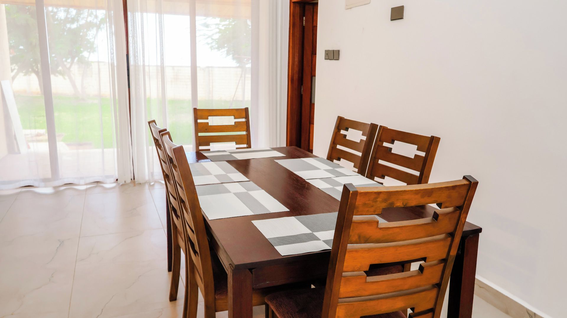 A dining area with a wooden table and chairs, a checkered table runner, and a view of a garden through large windows.