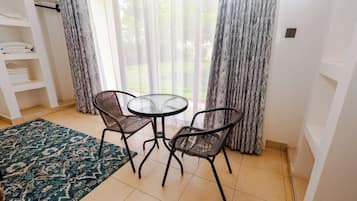 A room with a glass-top table and two wicker chairs, a blue patterned rug, and a white shelving unit with folded towels.