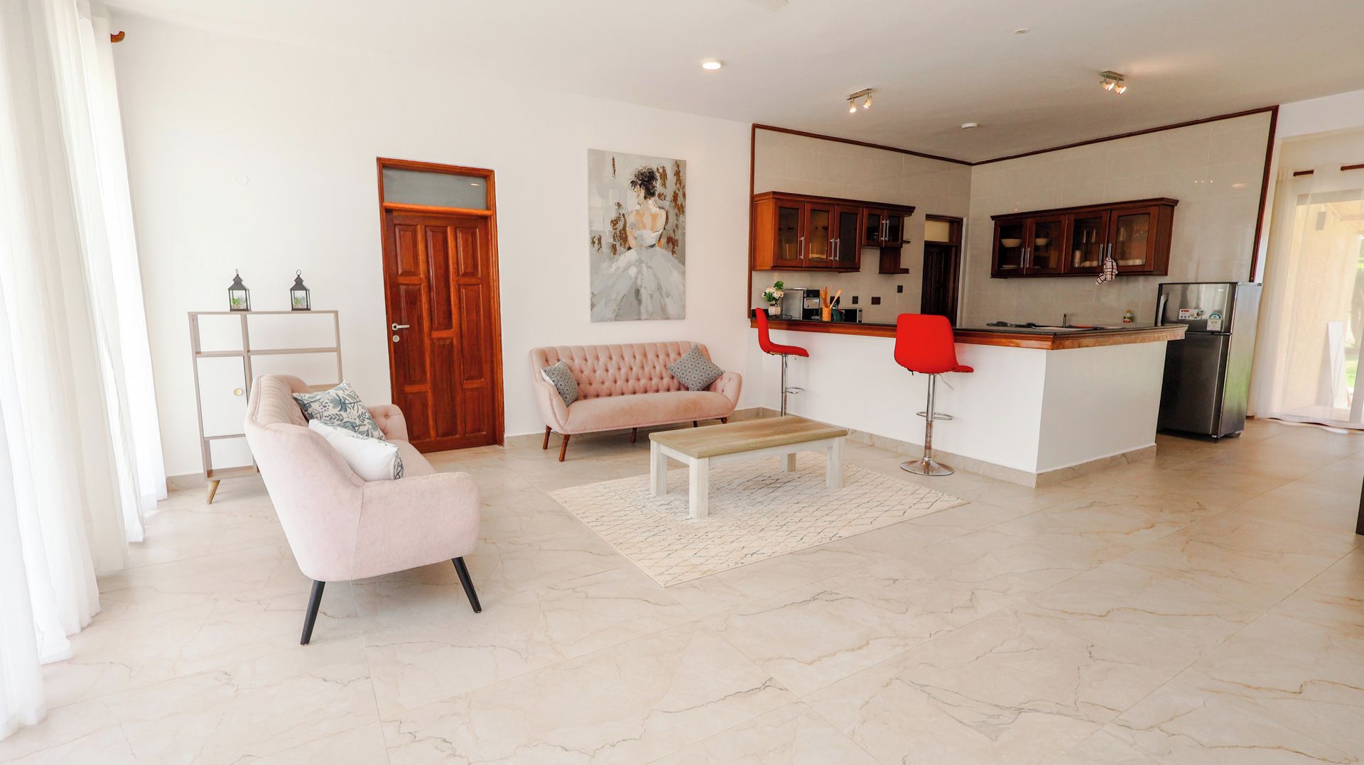 Una sala moderna con un sofá, una mesa de centro y un taburete rojo. Se observa una zona de cocina con armarios y un refrigerador.