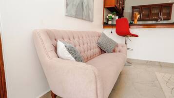 A pink sofa with patterned cushions in a room with a kitchen area in the background.