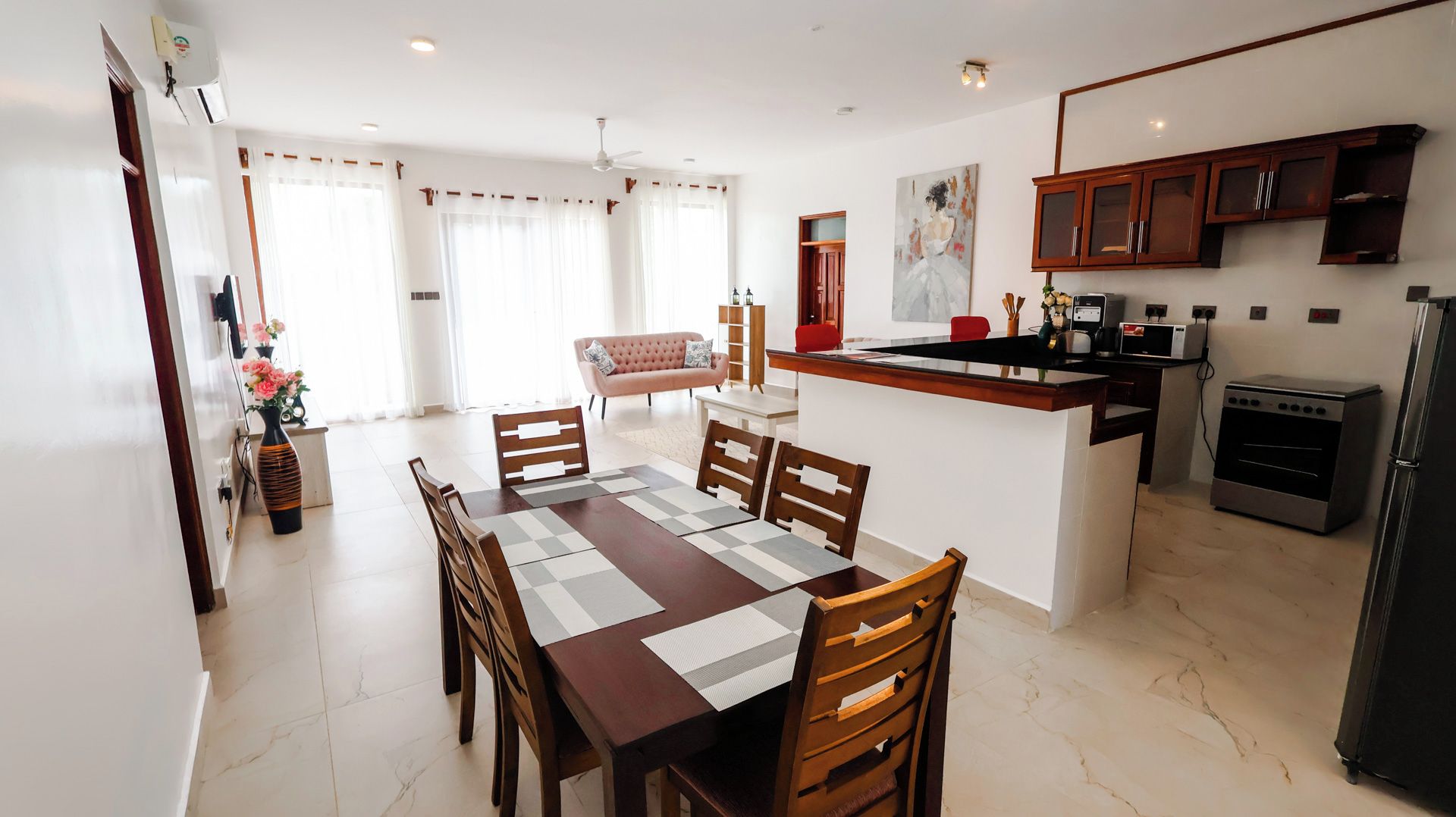 A modern kitchen with a built-in oven and refrigerator, a dining area with a wooden table and chairs, and a living space with a sofa and curtains.