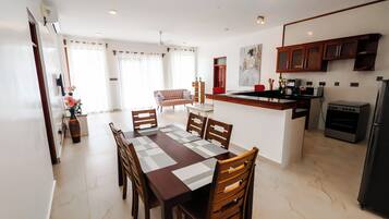 A modern kitchen with a built-in oven and refrigerator, a dining area with a wooden table and chairs, and a living space with a sofa and curtains.
