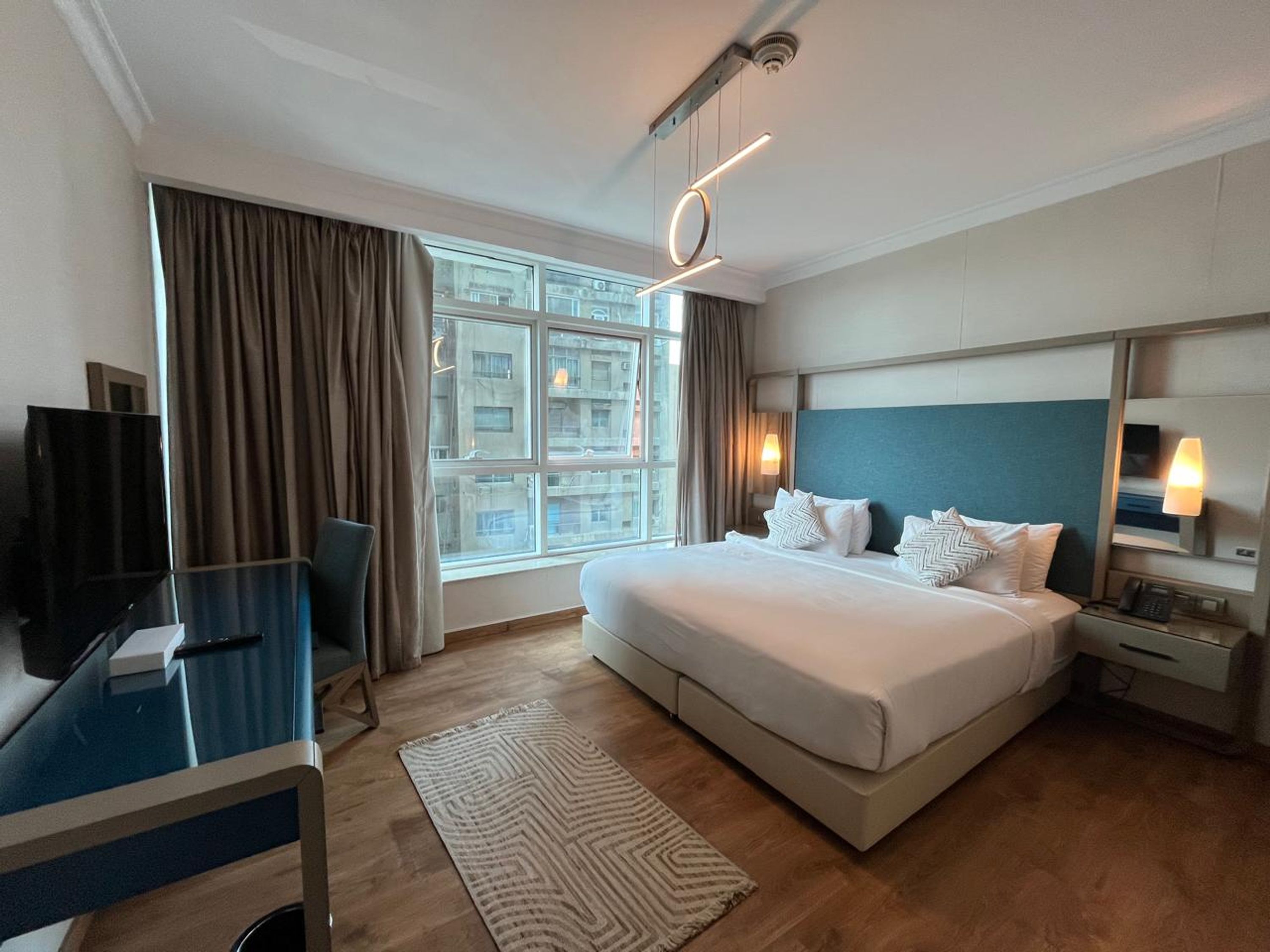 Habitación de hotel con una cama grande, un escritorio, un televisor, una ventana con cortinas y vistas a edificios.