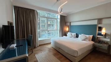 Habitación de hotel con una cama grande, un escritorio, un televisor, una ventana con cortinas y vistas a edificios.