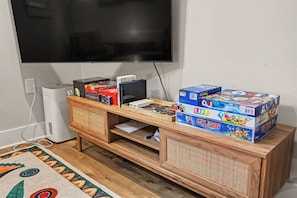 A room with a television mounted on the wall, a wooden entertainment unit with various board games, and a patterned rug on the floor. - The Social Canvas | Spacious & Cozy | Fire Pit, Blackstone, Game Room, Espresso Machine | Pet's OK! (Charlotte)