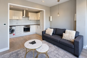 En moderne stue med en grå sofa, to hvide marmorborde og en vaskemaskine i det tilstødende køkkenområde.
