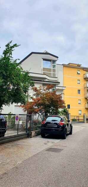 Uma área de estacionamento com um carro preto, um edifício com fachada branca e um edifício amarelo ao fundo.