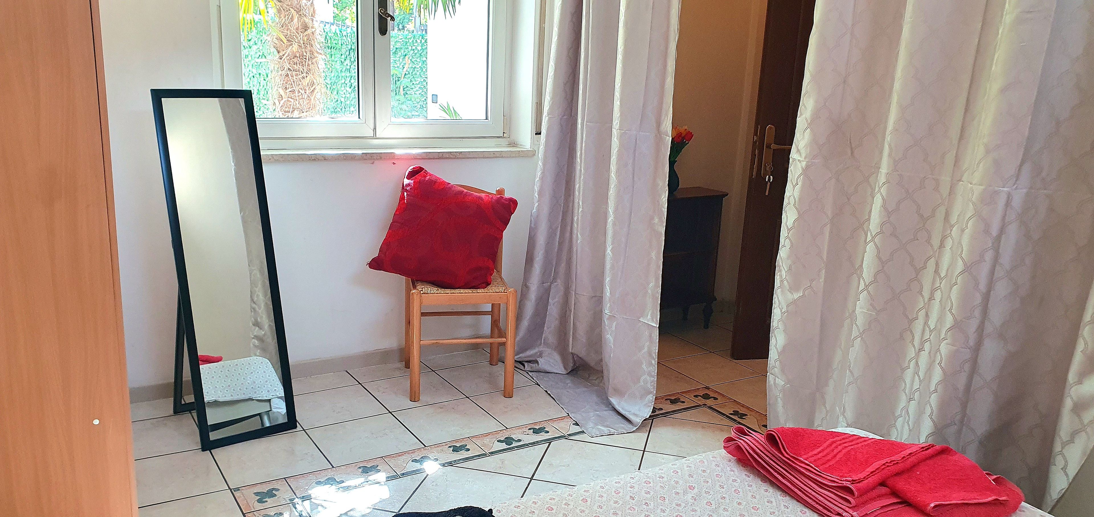 A room with a large mirror, a wooden chair with a red cushion, a window with curtains, and a tiled floor.