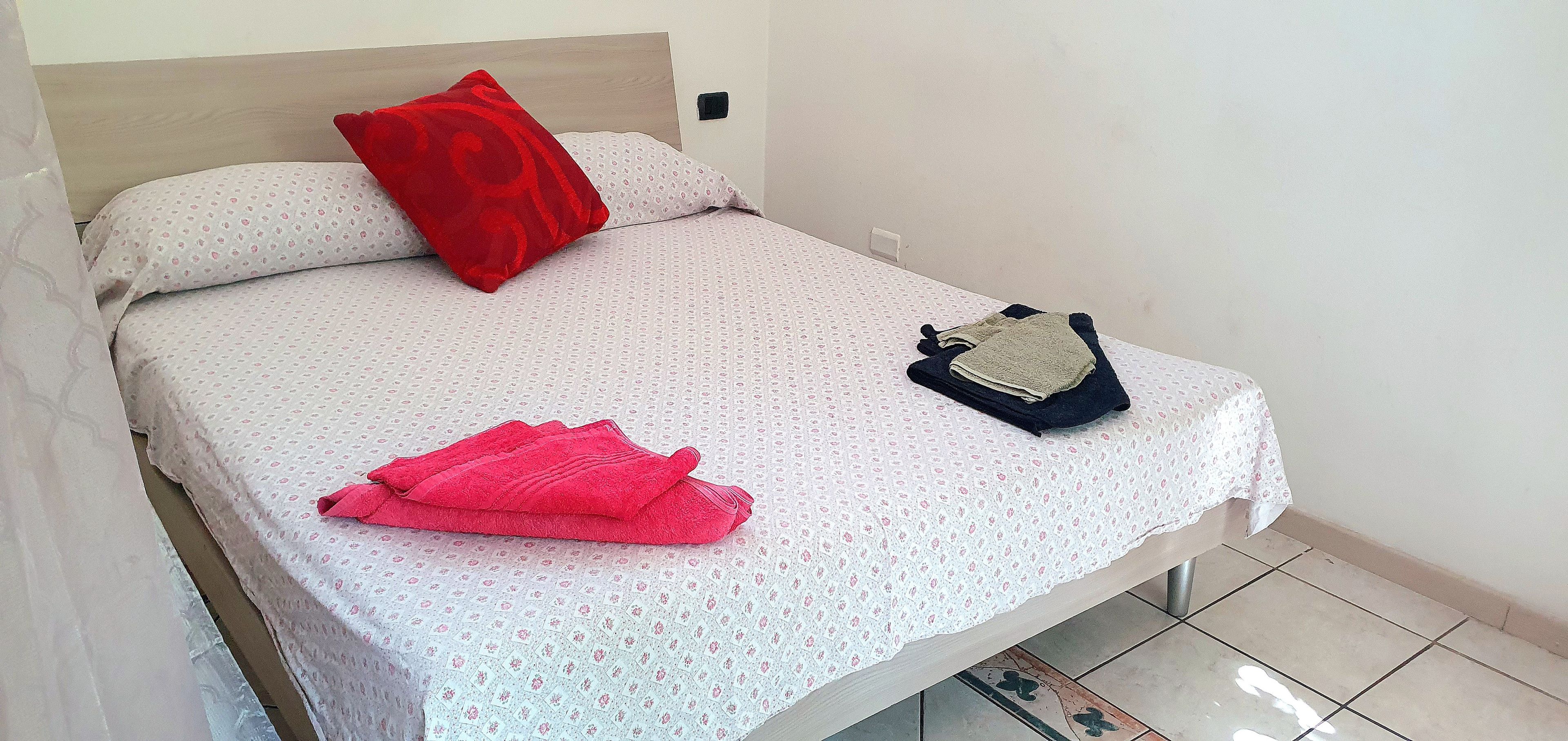 Een bed met een bedsprei met dessin, een rood kussen en een opgevouwen handdoek.