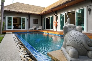 Una piscina con una estatua de piedra, techo de paja y vistas a una casa con ventanas y puerta grandes.
