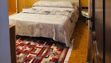 Un dormitorio con cabecera de madera, una cama con ropa de cama estampada, una alfombra roja y blanca, y un refrigerador.