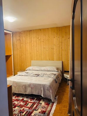 A bedroom with a wooden headboard, a bed with patterned bedding, a red and white rug, and a refrigerator. - Apartment 'Appartamento con due stanze Bolzano' (Bolzano)