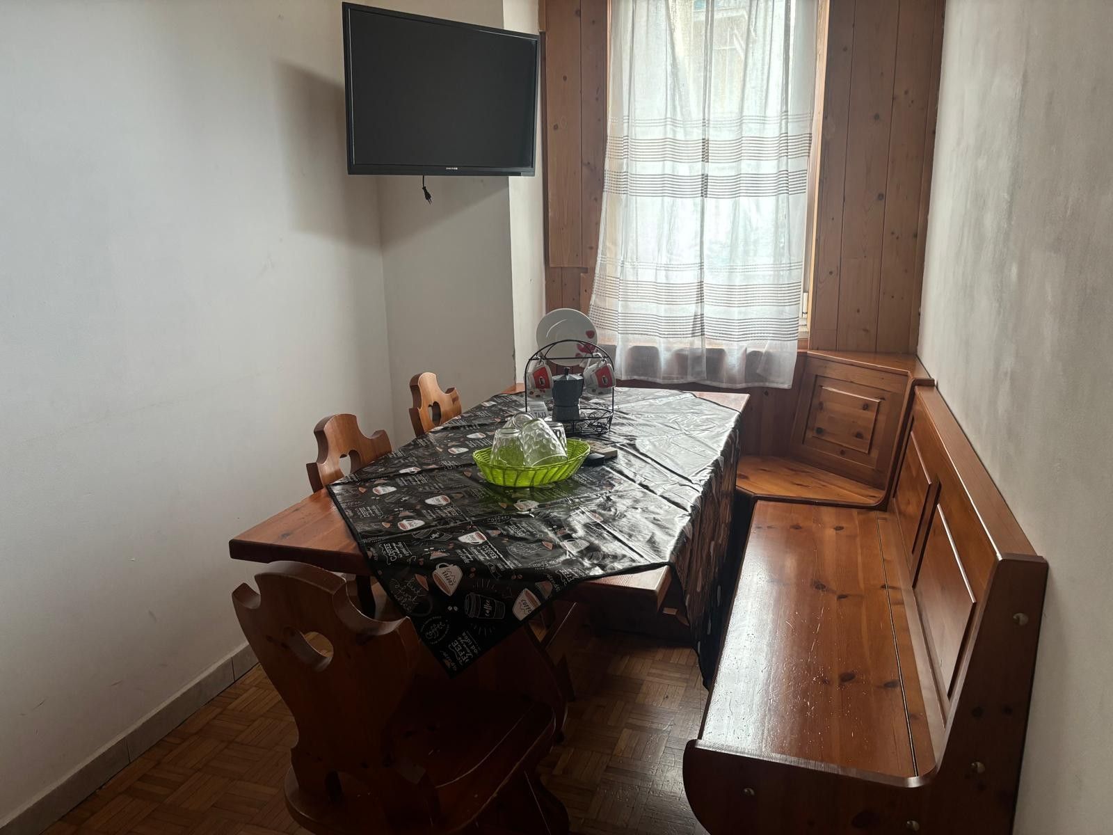 A dining area with a table, chairs, a television mounted on the wall, and a window with curtains.