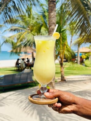 Eine Hand hält ein Glas mit einem gelben Getränk, einer Zitronenscheibe und einem Strohhalm. Im Hintergrund sind Palmen und ein Strand zu sehen.