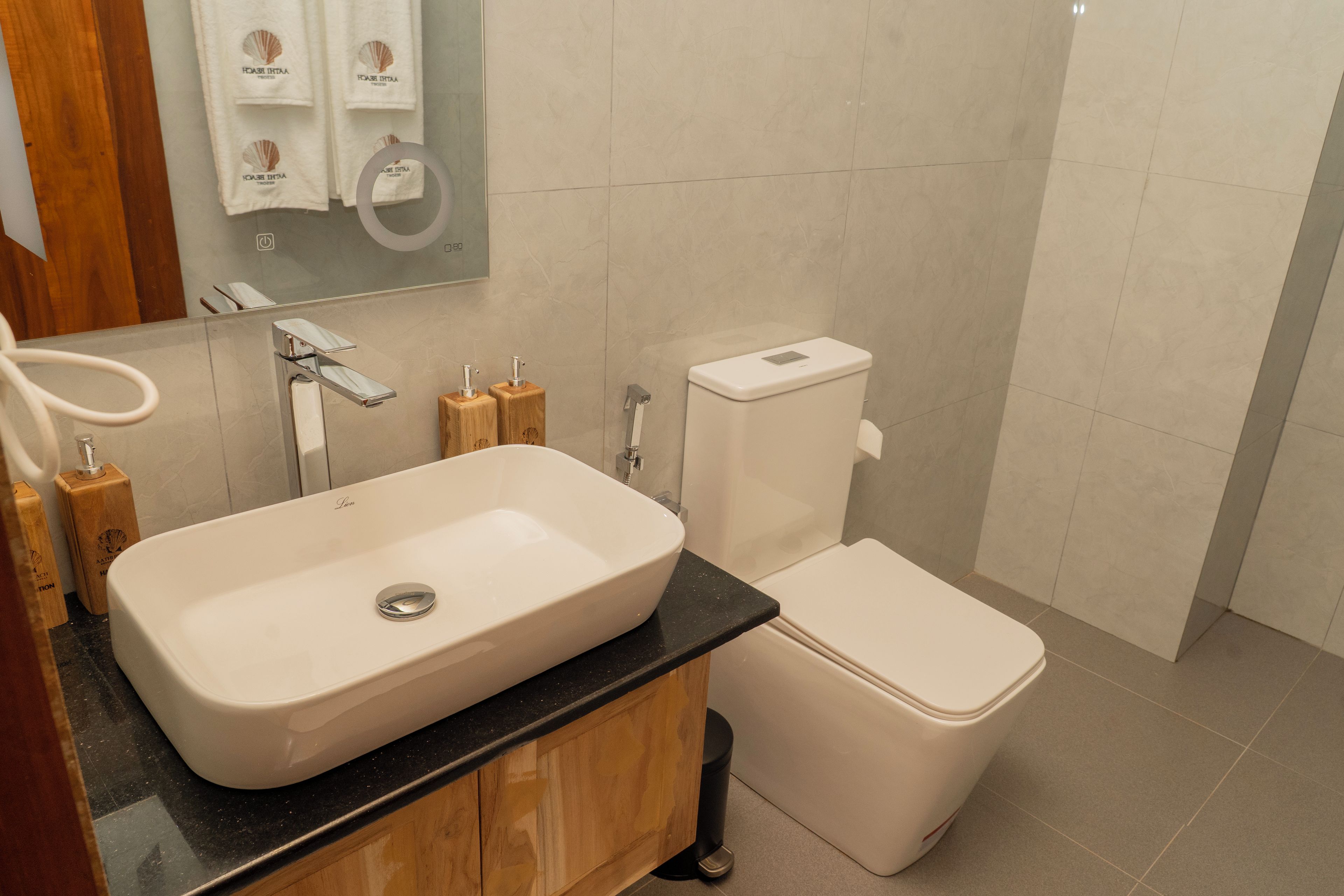 Une salle de bain avec un lavabo blanc, des toilettes et un miroir. On trouve des meubles en bois sous le lavabo.