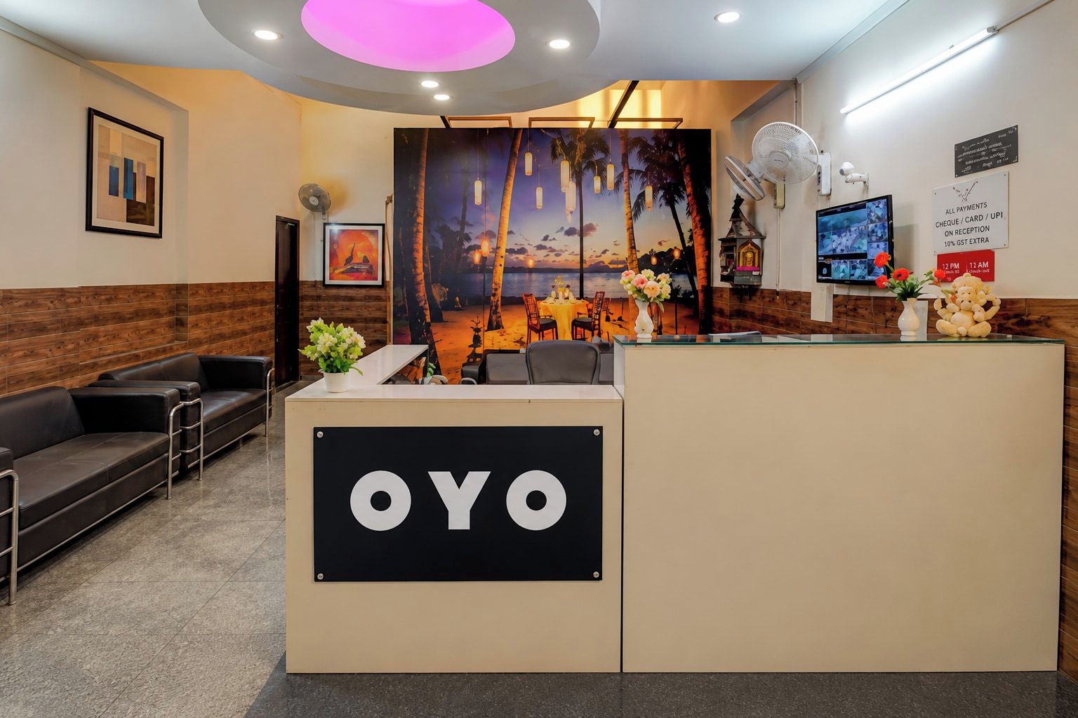 A reception area with a reception desk labeled 'OYO,' a seating area with a sofa, a wall-mounted fan, and a poster depicting a sunset scene.