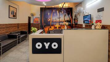 A reception area with a reception desk labeled 'OYO,' a seating area with a sofa, a wall-mounted fan, and a poster depicting a sunset scene.