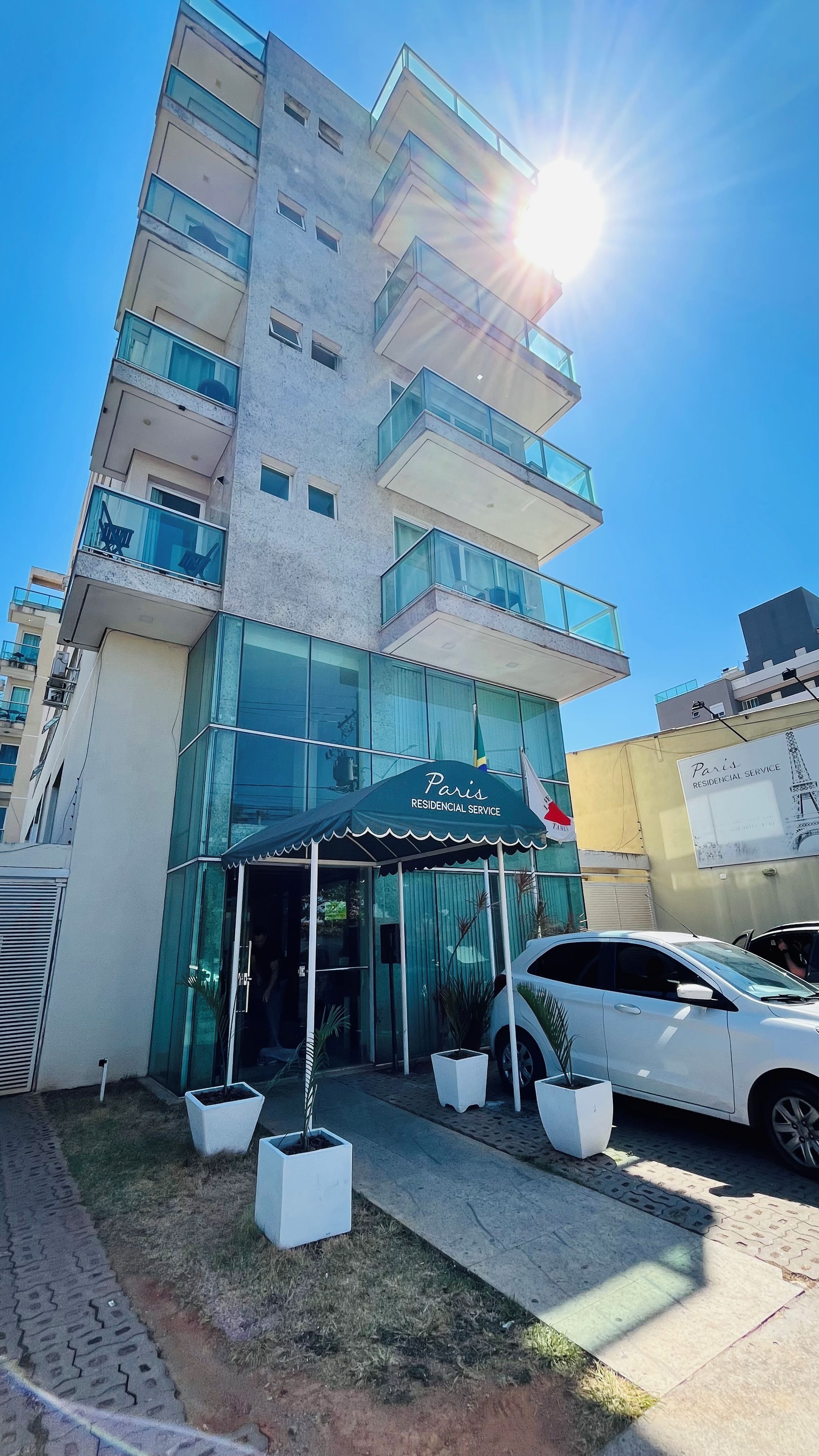 Un edificio de varios pisos con fachada de vidrio, un acceso cubierto y un coche blanco estacionado adelante.