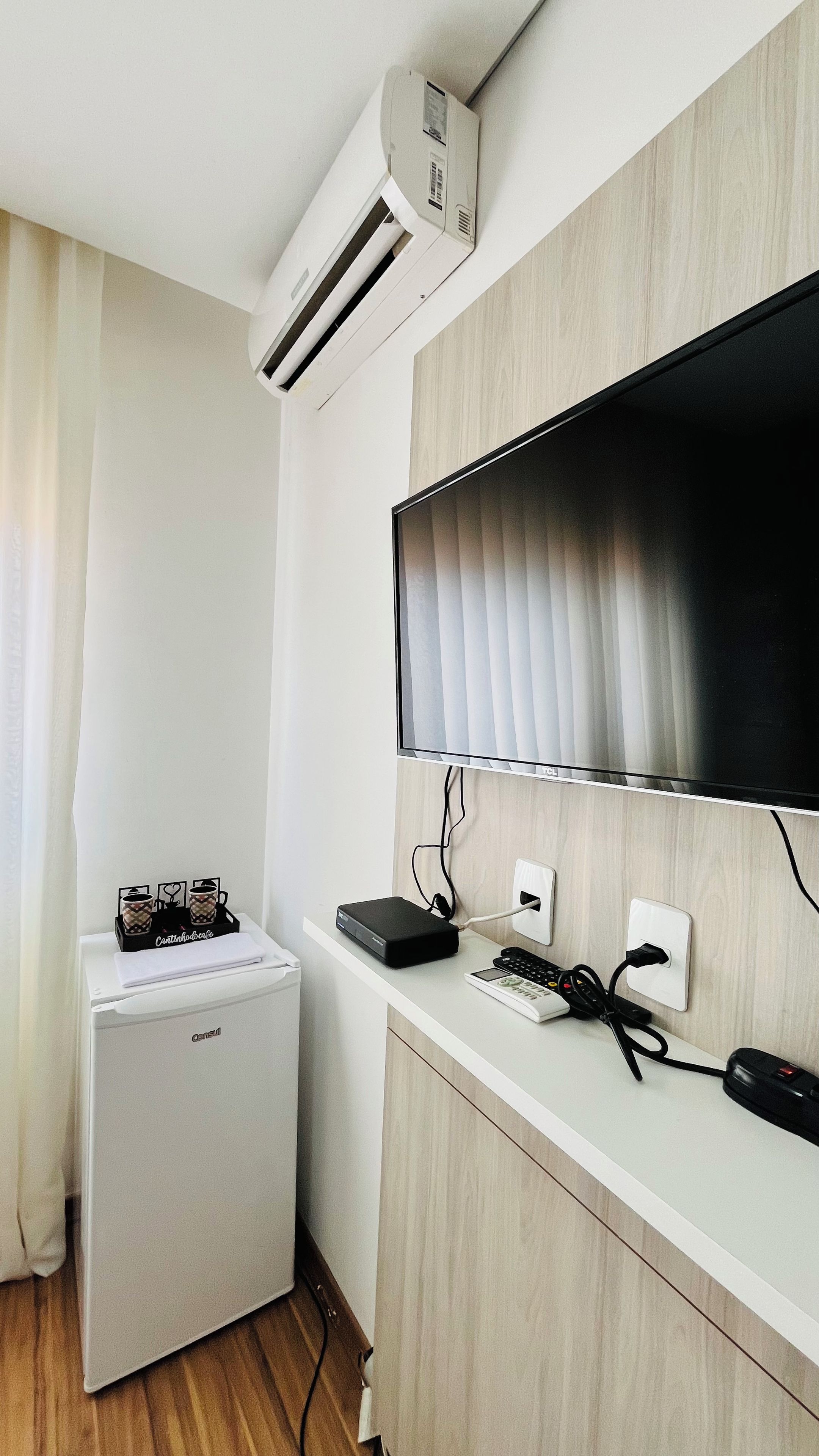 A compact room with a white mini-fridge, a flat-screen TV mounted on a wooden wall, and a small air conditioning unit above the fridge.