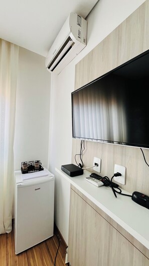 A compact room with a white mini-fridge, a flat-screen TV mounted on a wooden wall, and a small air conditioning unit above the fridge. - Apartment - Flat Belotto 01 __ 15 minutes from Confins Airport (Lagoa Santa)