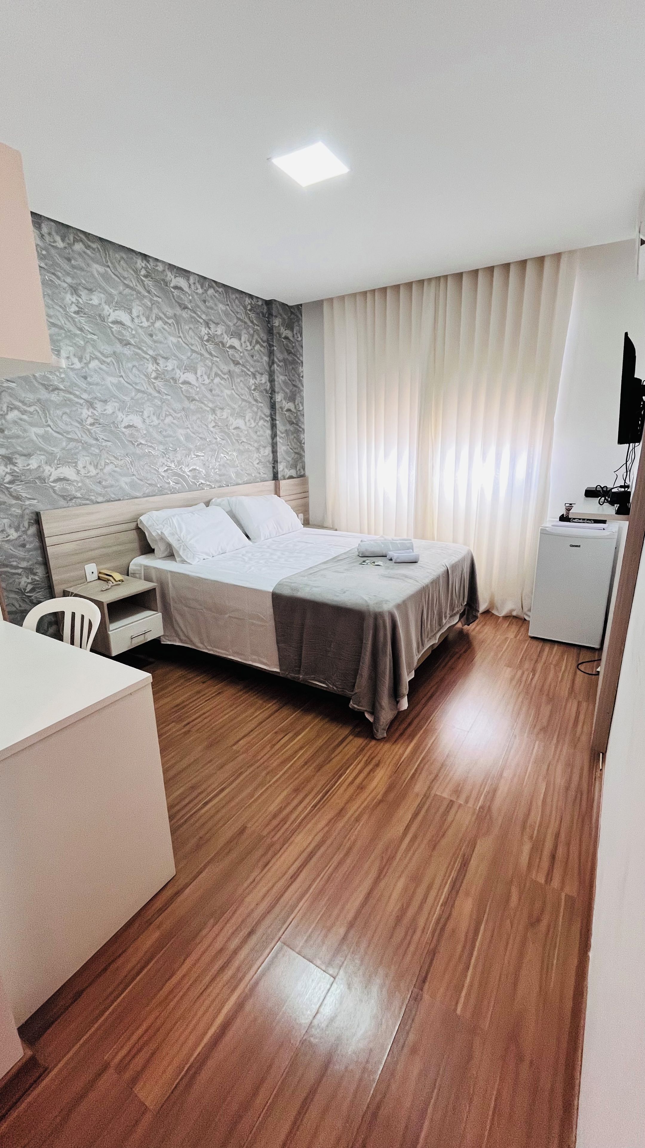 A hotel room with a bed, bedside table, chair, desk, and a small refrigerator.