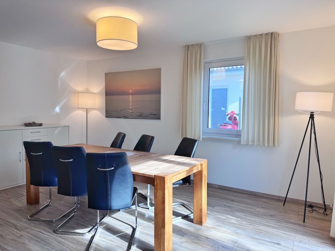 Habitación de hotel con mesa de comedor de madera, cuatro sillas, lámpara de pie, lámpara de suelo, ventana con cortinas y un cuadro de un atardecer.