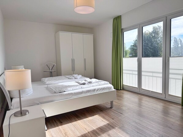 Un dormitorio moderno con cama, mesita de noche, lámpara, armario y ventanas amplias.