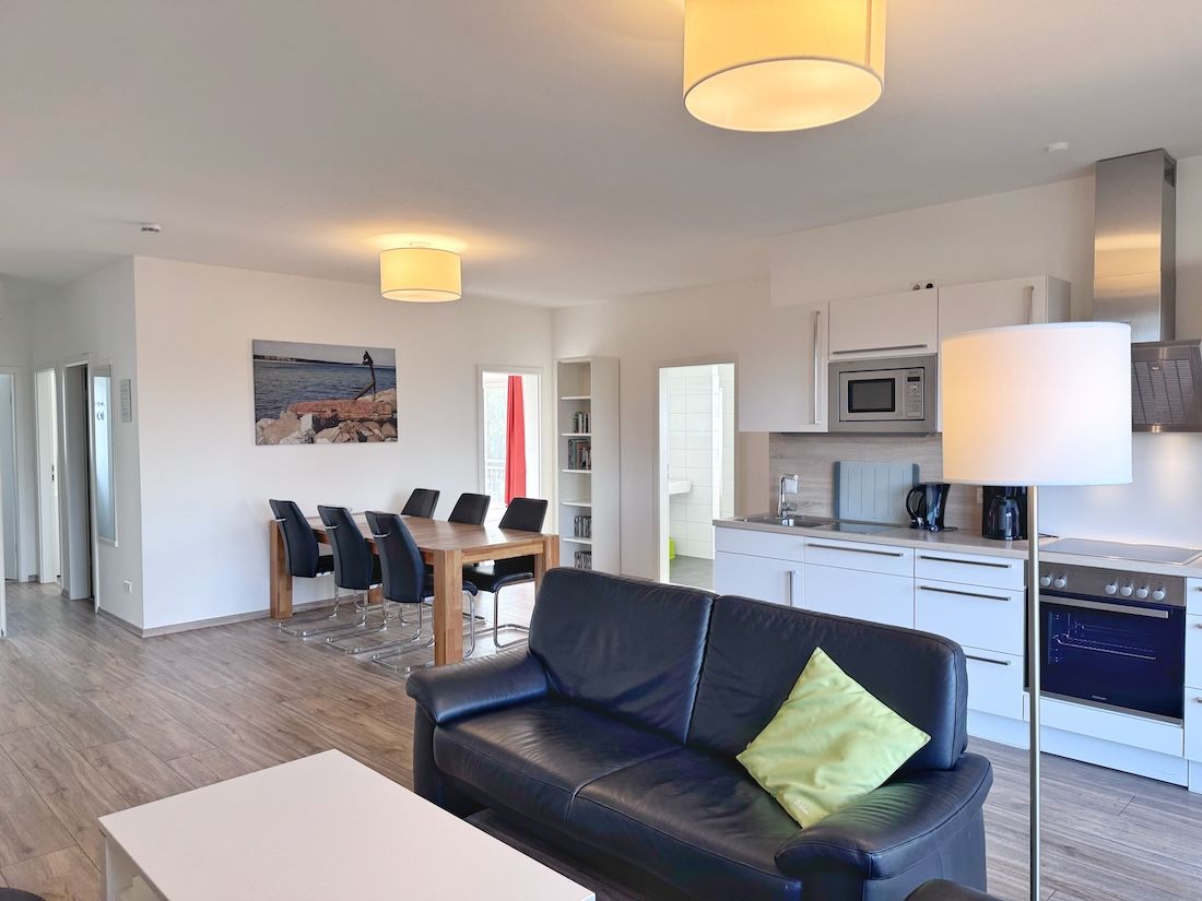 A modern living room with a black leather sofa, a dining table with chairs, a kitchen area with a microwave, stove, and cabinets, and a painting on the wall.