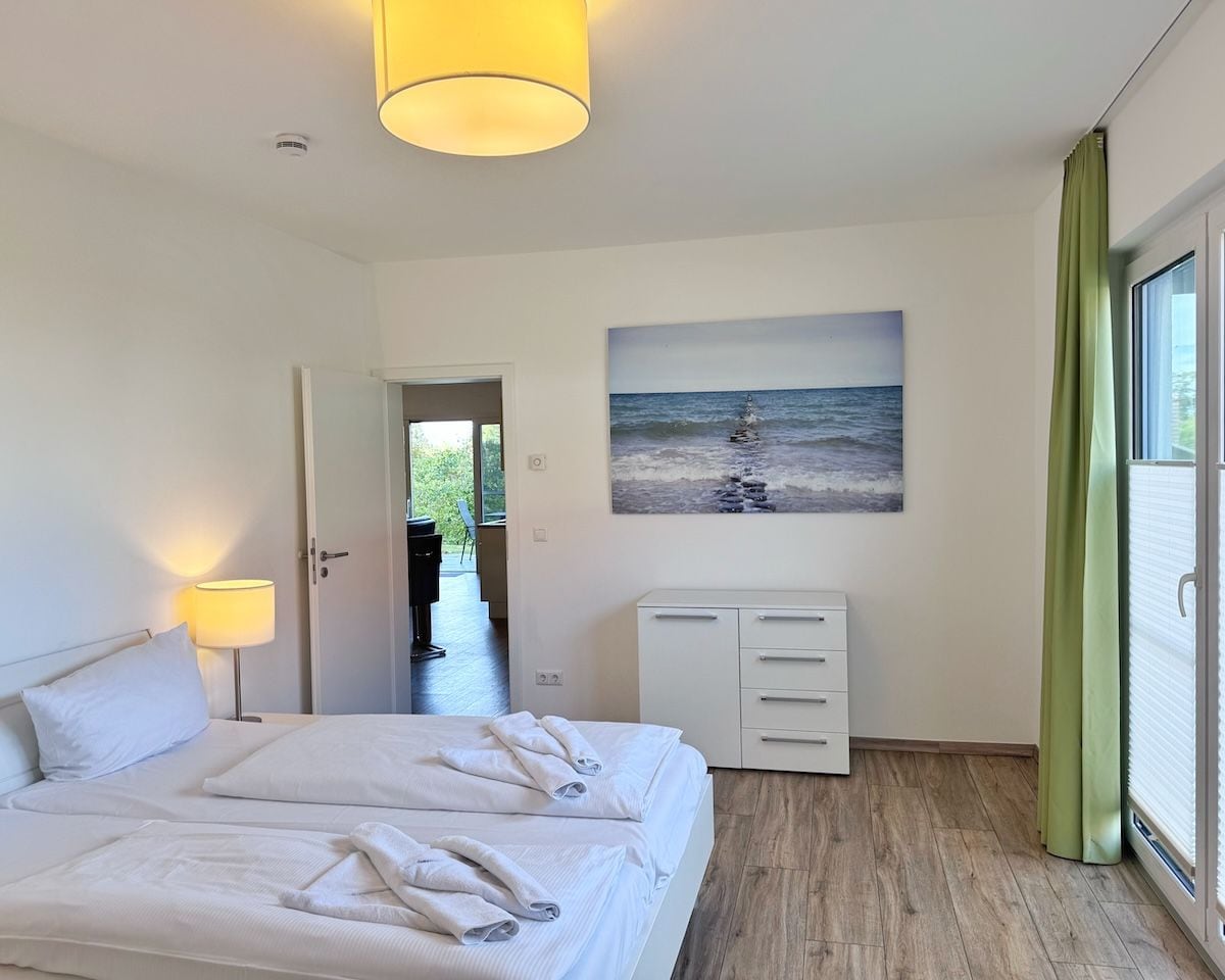 Une chambre d’hôtel avec un grand lit, un tableau représentant une scène de plage, un bureau avec des tiroirs et une vue par la fenêtre.