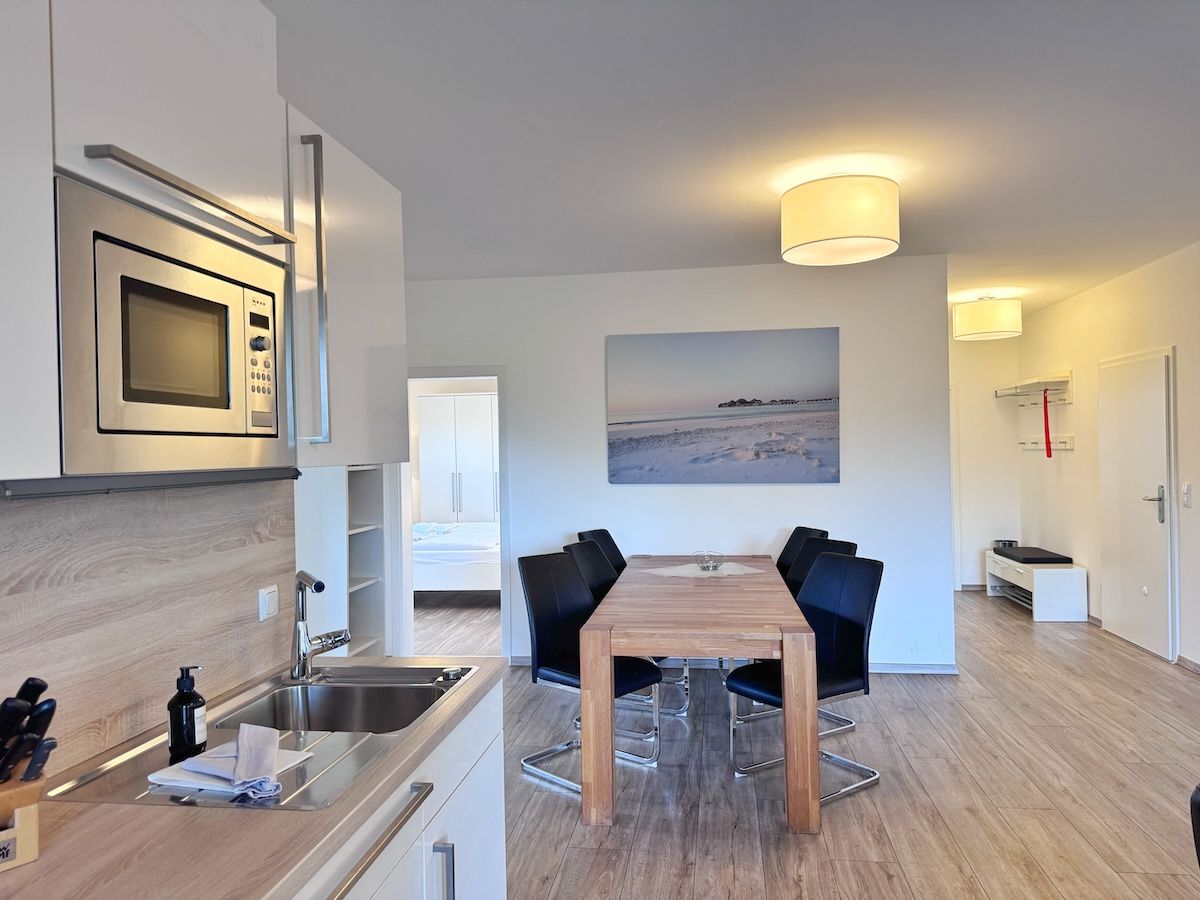 Une cuisine moderne équipée d’un four à micro-ondes encastré, un coin repas avec une table en bois et des chaises noires, et un tableau accroché au mur.