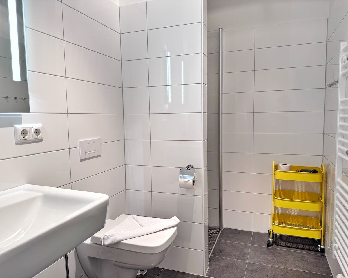 Une salle de bain avec des carreaux blancs, un lavabo blanc, des toilettes, une cabine de douche et un chariot de nettoyage jaune.