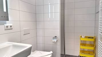 Une salle de bain avec des carreaux blancs, un lavabo blanc, des toilettes, une cabine de douche et un chariot de nettoyage jaune.