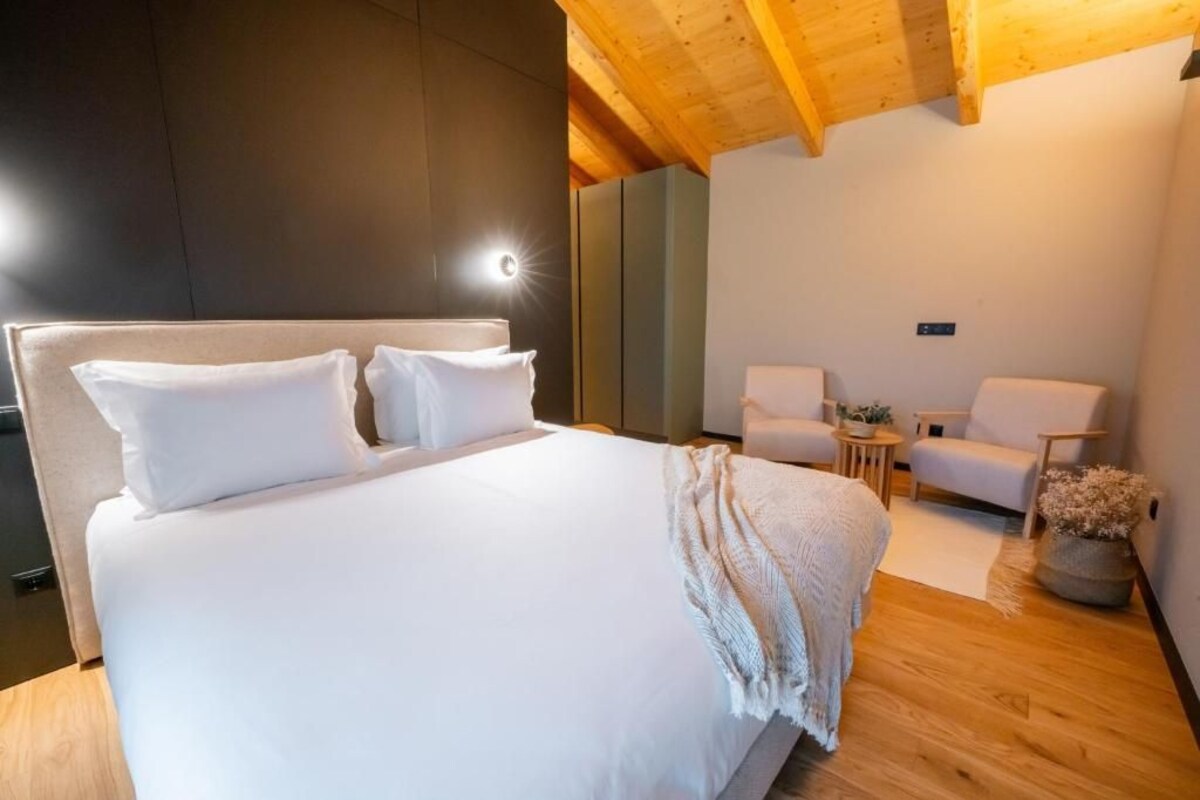A modern bedroom with a large bed, two armchairs, a small table, and a wooden ceiling.